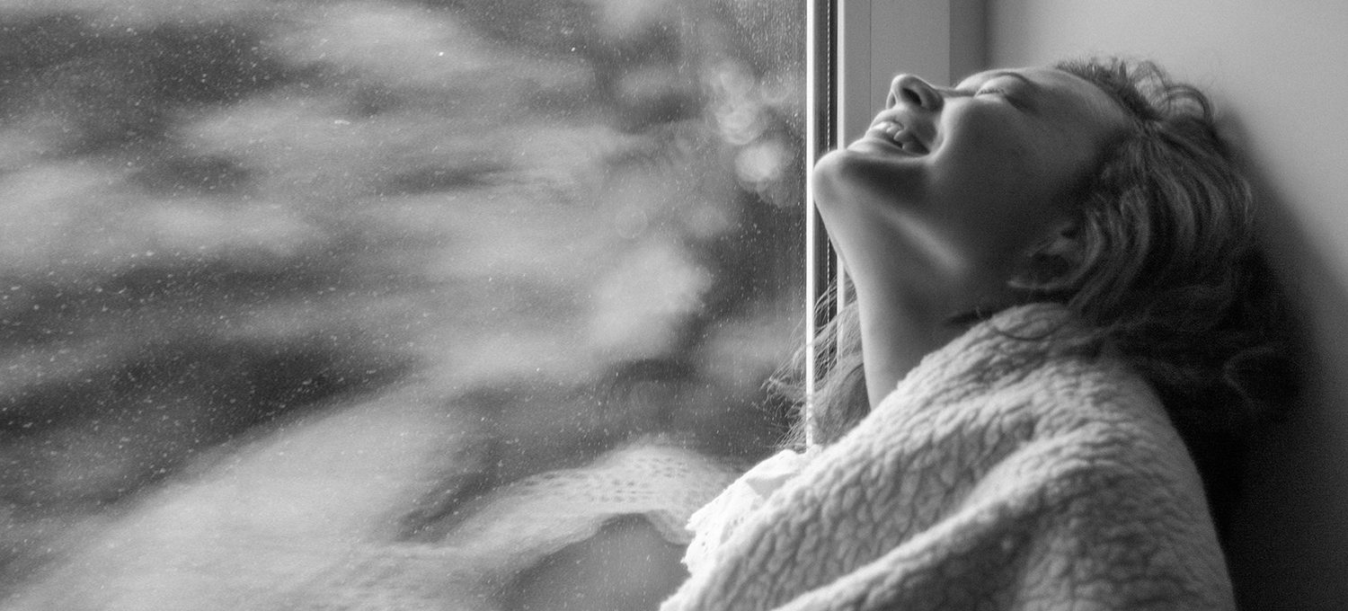 Frau in schwarz weiss sitzt mit einer dicken decke im Fenster rahmen und lacht