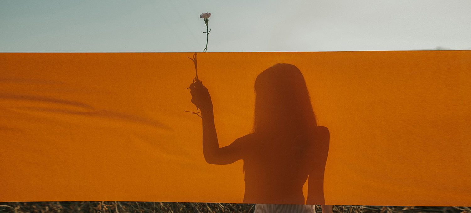 Silhouette einer Frau hinter einem orangen Leintuch. Sie hält eine Blume in der Hand welche oben heraus schaut.