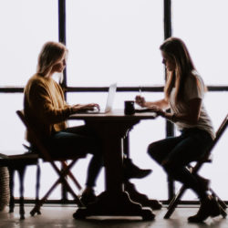 zwei Frauen sitzen sich in einem Loft an einem Tisch gegenüber und arbeiten. Gegenlicht durch das Fenster vor dem sie sitzen.