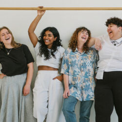 4 Frauen mit unterschiedlichen Hautfarben und Kleidergrößen stehen lachend vor eine weißen Wand